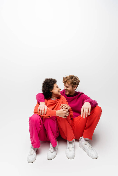 positive african american woman sitting with happy man in magenta color sweatshirt on white  - Photo, Image