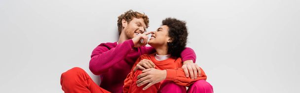 happy man toughing nose of african american woman in magenta color clothes on grey, banner  - Foto, Imagen