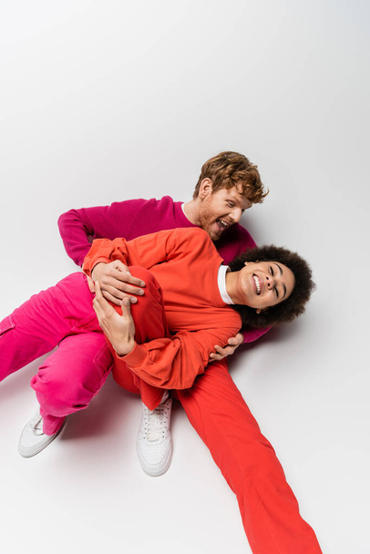 overhead view of happy redhead man in magenta color sweatshirt hugging cheerful african american woman on white  - Photo, Image