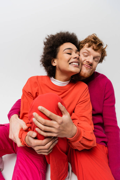 happy redhead man in magenta color sweatshirt hugging cheerful african american woman on grey  - Foto, immagini
