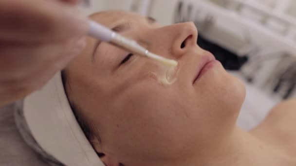 Enzimterápia. Kozmetikus alkalmazása hidratáló orvosi hámlasztó krém maszk fiatal nő arcát ecsetekkel. Lány bőr eljárás spa. Arcplasztikai kezelés. Természetes kozmetikai termékek - Felvétel, videó
