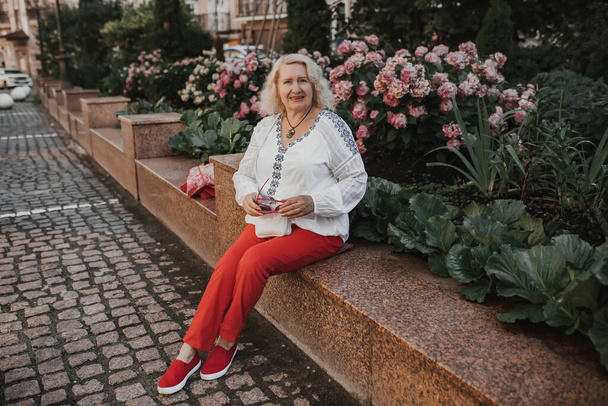 An elderly woman sits near azaleas in the city while traveling or walking. - Fotoğraf, Görsel
