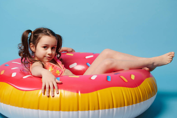 Lovely little child girl with two ponytails, wearing a stylish swimsuit, looking at camera while lying on an inflatable donut swimming ring, a colorful lifebuoy, isolated over blue color background. - Photo, Image