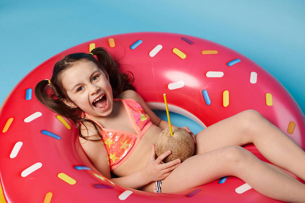 Вид сверху очаровательная маленькая девочка в купальнике, с кокосовым напитком, улыбающаяся, глядя в камеру, расслабляющаяся на плавающем пончике плавательном кольце, изолированный синий фон. Летние развлечения и концепция пляжа - Фото, изображение