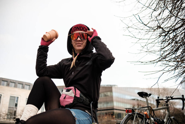 Bike courier woman is sitting outside having a cup of coffee and looking at camera wearing sunglasses. She has a bike behind her. Job break concept. - 写真・画像