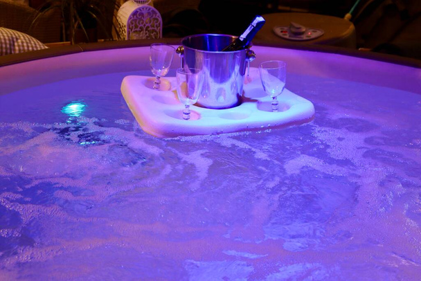Botella de champán en cubo de hielo y 4 vasos en un flotador en un baño jacuzzi burbujeante con luces púrpuras bajo el agua - Foto, imagen