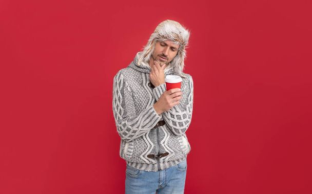 pondering grizzled guy in sweater and earflap hat drink coffee on red background, lifestyle. - Foto, Imagen