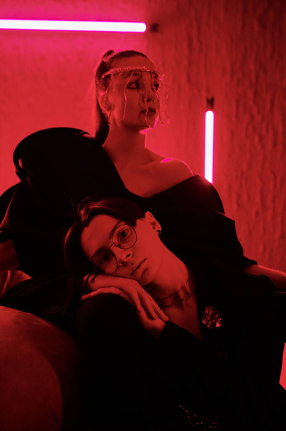 Affectionate teenage couple in stage apparel relaxing in front of camera at vogue ball event in modern night club lit with red neon light - Foto, Bild