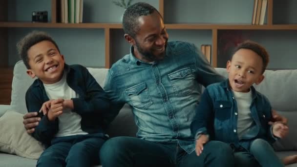 Afro-Amerikaanse volwassen vader omhelst knuffelen praten lachen met twee kleine jongens kleuterschool kinderen zonen die plezier gesprek knuffelen zitten op de bank. Liefdevolle vader praat graag met kinderen op de bank - Video