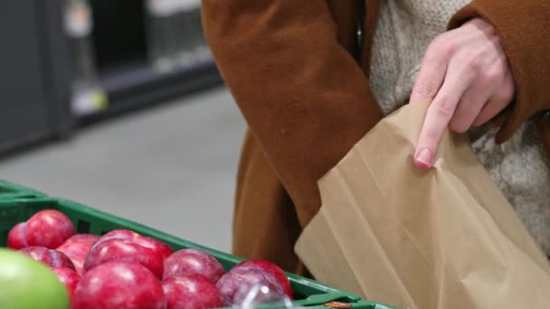 Young caucasian unrecognizable man in a brown coat and knitted sweater takes red round plums from the counter, examines and puts in a paper bag, close-up side view.Offline shopping concept. - Filmagem, Vídeo