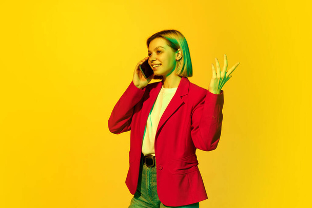 Half-length portrait of young emotional girl with blonde hair wearing red jacket talking on smartphone over yellow background. Concept of youth, student college life, business and education. - Photo, Image