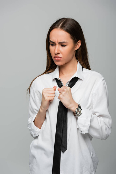 angespannte und brünette Frau in weißem Hemd und Krawatte mit Blick auf die Armbanduhr vereinzelt auf grau - Foto, Bild