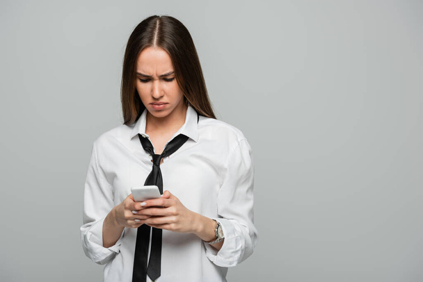 νεαρή γυναίκα με λευκό πουκάμισο και μηνύματα γραβάτα στο smartphone απομονώνονται σε γκρι  - Φωτογραφία, εικόνα