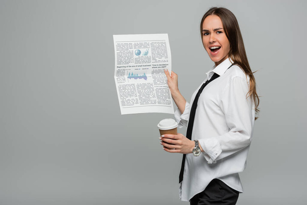 užaslá mladá žena v bílé košili s kravatou drží noviny a papírový šálek izolované na šedé  - Fotografie, Obrázek