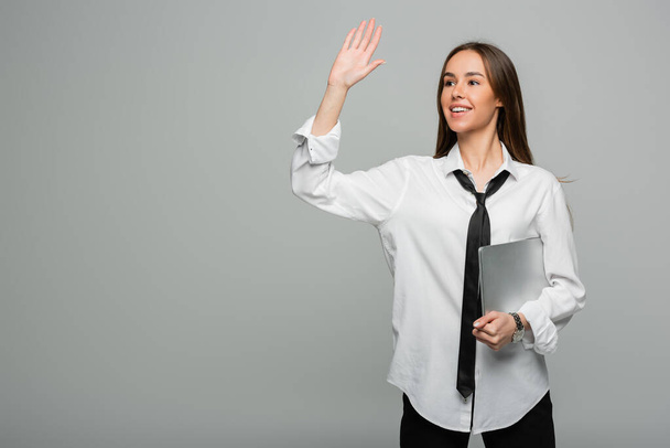 happy woman in white shirt with tie holding laptop while waving hand isolated on grey  - Photo, Image