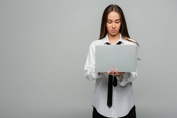 fiatal barna nő fehér ing nyakkendő segítségével laptop elszigetelt szürke  - Fotó, kép