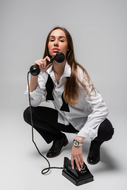 young woman in white shirt with tie and pants holding retro telephone while sitting on grey  - Zdjęcie, obraz