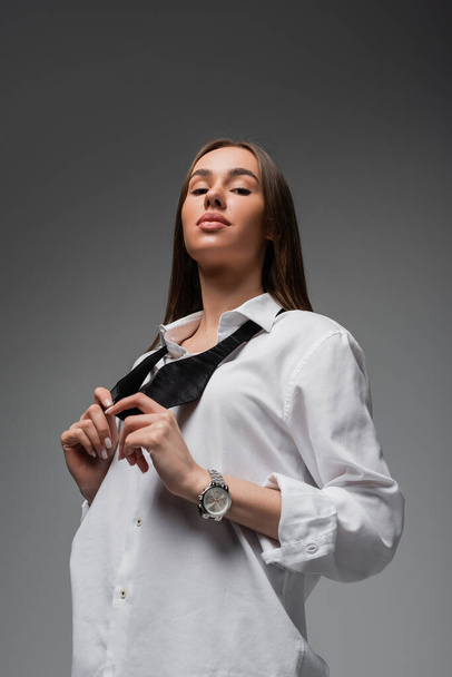 портрет уверенной женщины в белой рубашке, регулирующей черный галстук при взгляде на камеру, изолированную на сером, концепция гендерного равенства  - Фото, изображение