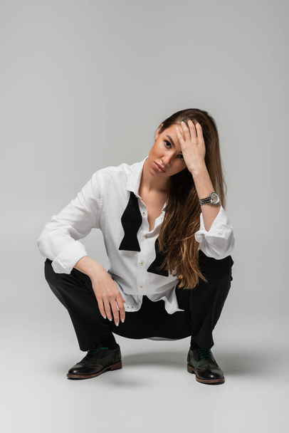 full length of brunette woman in shirt with tie and black pants sitting on grey, gender equality concept  - Photo, image