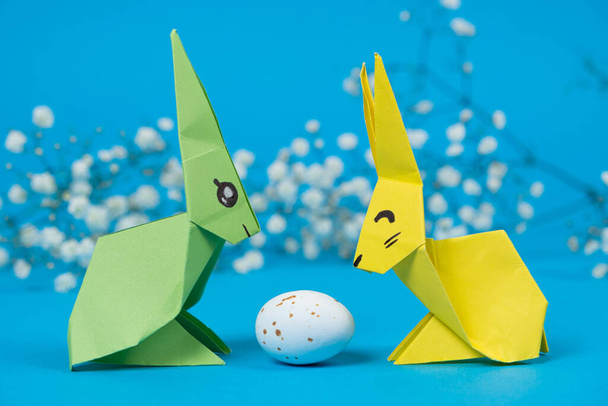 Due coniglietti di origami di carta e un uovo di Pasqua su sfondo blu. Artigianato con le proprie mani per Pasqua. - Foto, immagini