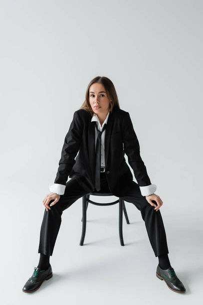 full length of young brunette woman in black suit with tie sitting on wooden chair on grey  - Foto, Imagem