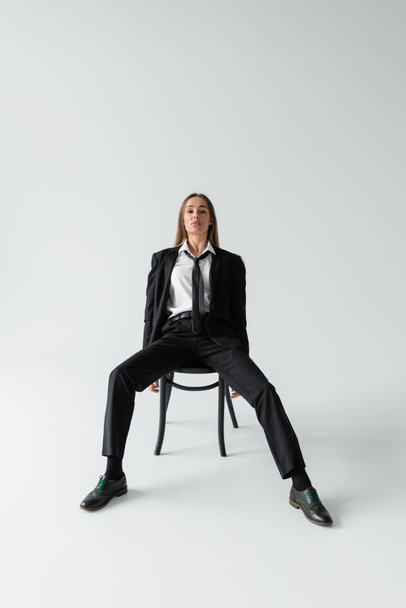 full length of young brunette woman in black suit with tie sitting on wooden chair while looking at camera on grey  - Fotoğraf, Görsel
