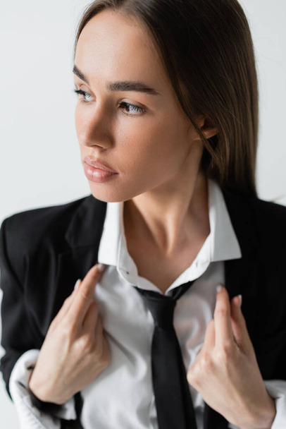 portrait of pretty and brunette woman in black suit with tie looking away isolated on grey  - Photo, image