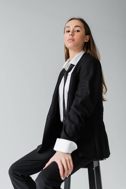 young brunette woman with long hair sitting in black suit with tie and looking at camera isolated on grey  - Fotó, kép