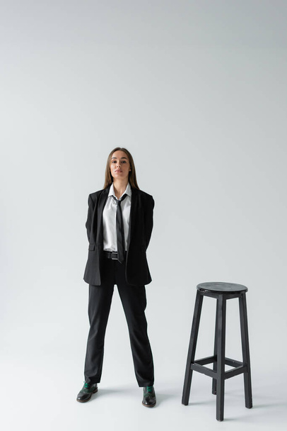 полная длина уверенной женщины в черном костюме с галстуком, стоящим рядом с высоким стулом на сером  - Фото, изображение