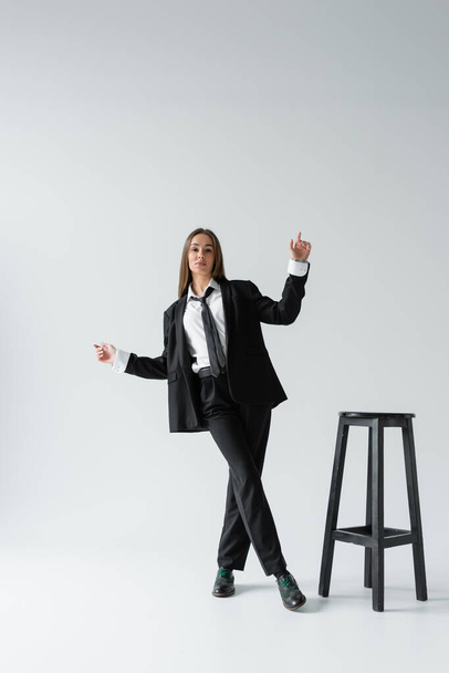 πλήρες μήκος της νεαρής μελαχρινής γυναίκας σε μαύρο κοστούμι με γραβάτα ποζάρει κοντά σε ψηλή καρέκλα σε γκρι  - Φωτογραφία, εικόνα