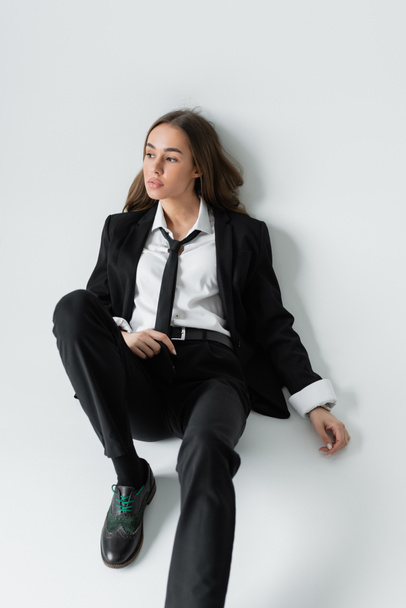 pełna długość zamyślony kobieta w czarny formalny odzież z krawat siedzi na szary tło  - Zdjęcie, obraz