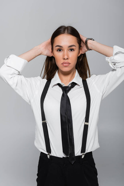brunette young woman in black pants and suspenders looking at camera isolated on grey  - Foto, Bild