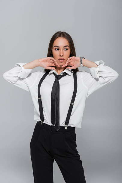 brunette young woman in black pants and suspenders looking away isolated on grey  - Photo, Image