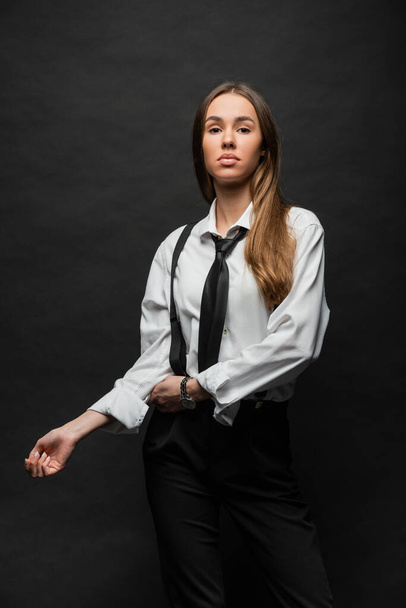 brunette woman with long hair standing in formal wear with suspenders while posing on black  - Zdjęcie, obraz
