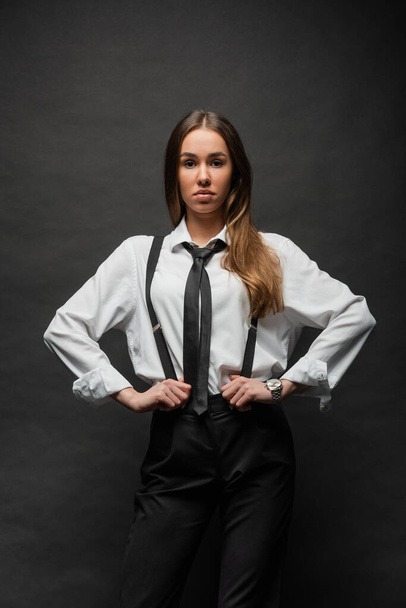 brunette woman with long hair standing in formal wear with hands on hips on black  - Zdjęcie, obraz
