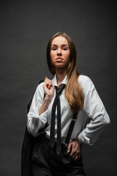 brunette woman with long hair standing in formal wear with suspenders while holding blazer on black  - 写真・画像