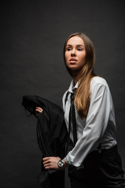 young brunette woman with long hair standing in suit while holding blazer on black background  - Foto, Bild