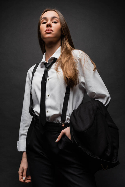 confident woman with long hair standing in suit with hand in pocket while holding blazer on black  - Fotografie, Obrázek