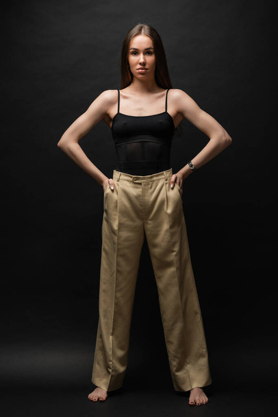 полная длина мускулистая женщина в верхней и бежевые брюки позируют с руками в карманах на черном фоне  - Фото, изображение