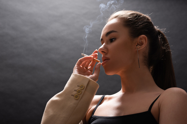 nízký úhel pohledu na mladé brunetky žena v béžové sako kouření cigarety na černém pozadí  - Fotografie, Obrázek