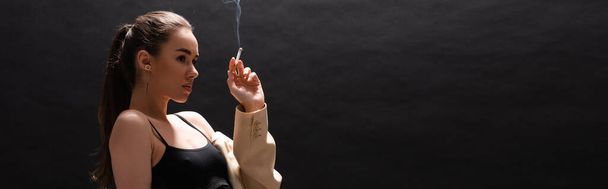 young brunette woman in beige blazer holding cigarette while looking away on black background, banner  - Photo, Image