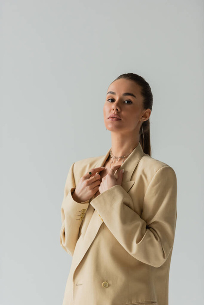 brunette young woman in beige jacket posing while looking at camera isolated on grey  - Photo, Image