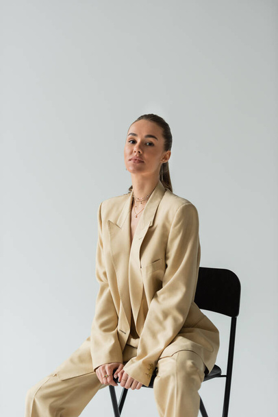 morena mujer joven en traje de moda beige sentado en silla aislada en gris  - Foto, imagen