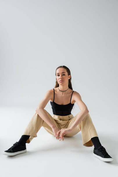 full length of pretty model in beige pants and black top looking at camera while sitting on grey  - Photo, Image