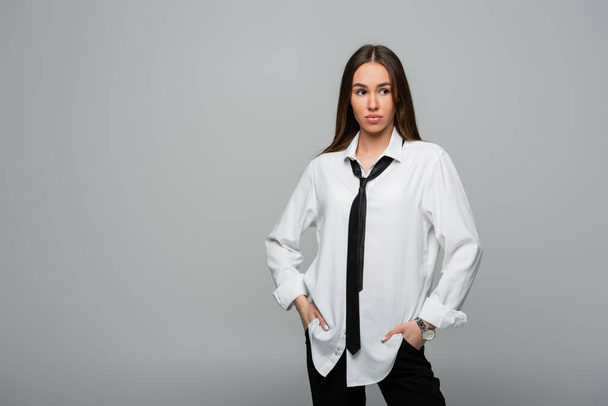young and brunette woman in white shirt with tie posing with hands in pockets isolated on gray  - Valokuva, kuva