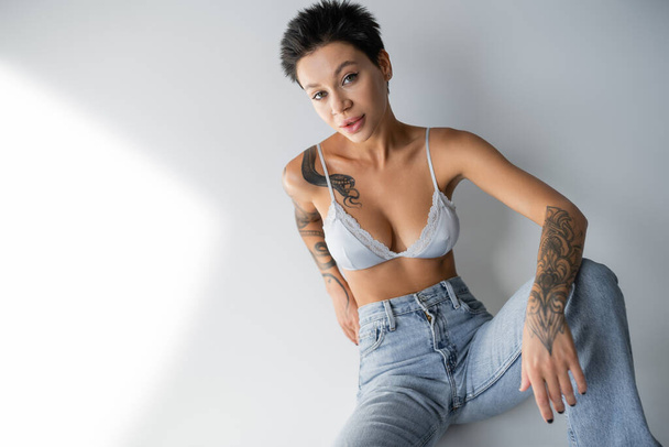 brunette woman with sexy tattooed body sitting in jeans and bra on grey background - Fotó, kép