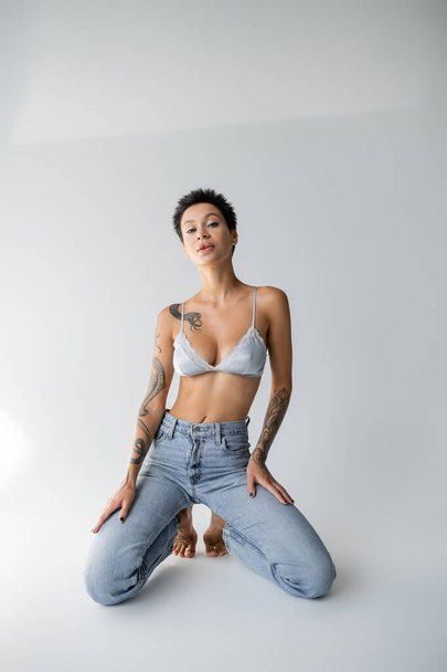 sensual brunette woman in satin bralette and jeans posing on knees on grey background - Photo, image