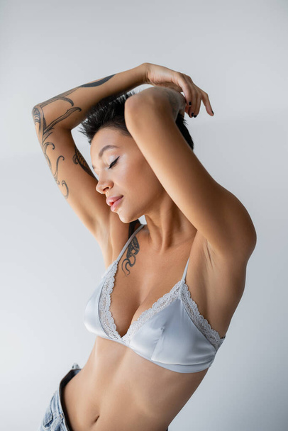 sensual tattooed woman in silk bralette posing with closed eyes and hands above head isolated on grey - Photo, Image