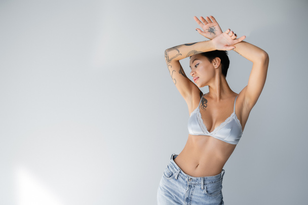 young and slender woman in satin bra and jeans posing with tattooed hands above head on grey background - Valokuva, kuva