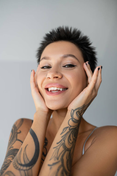 portrait of cheerful tattooed woman with short hair and piercing holding hands near face on grey background - Photo, image